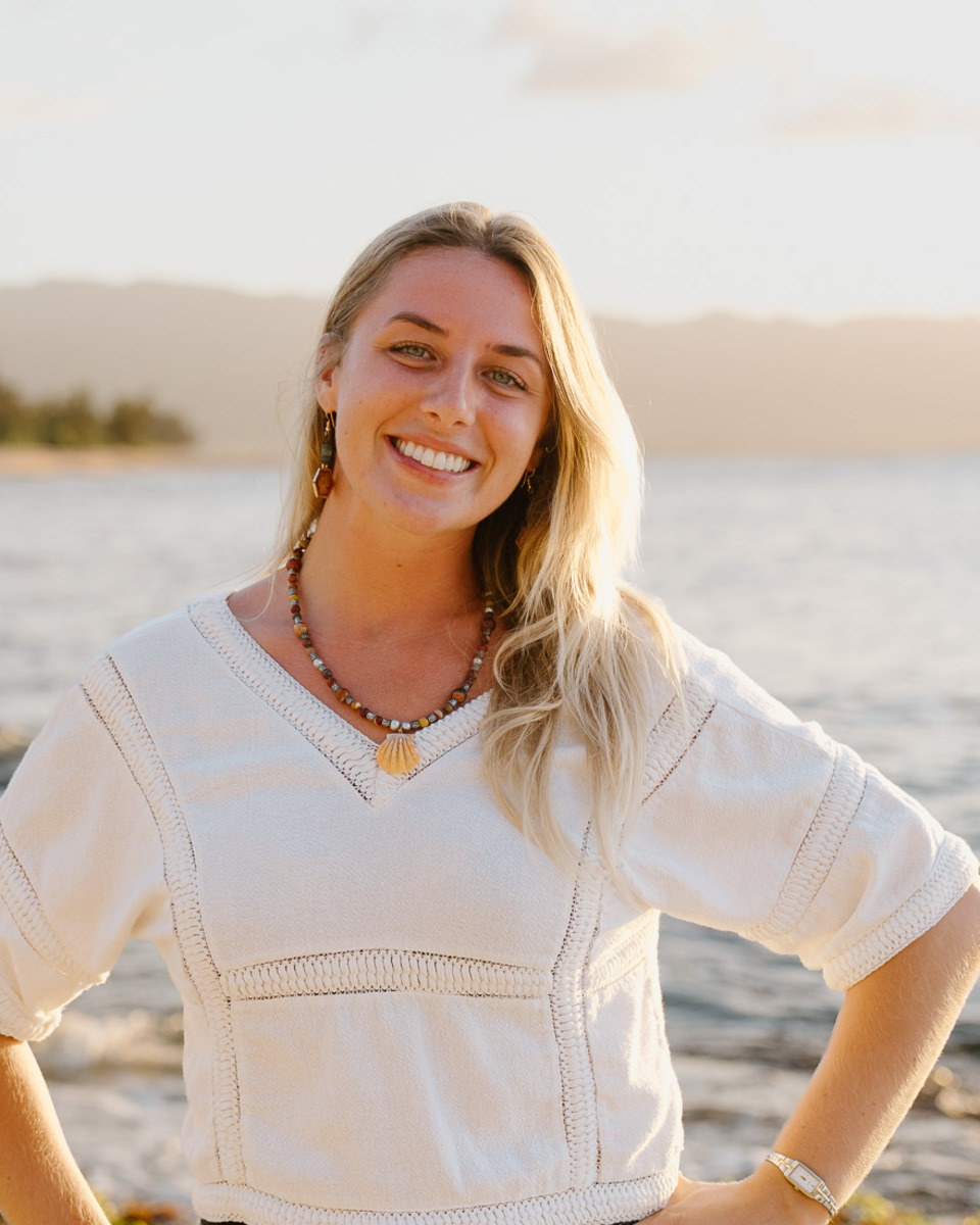 Lindsey on the beach at golden hour, in a cream top, hands on hips, smiling.