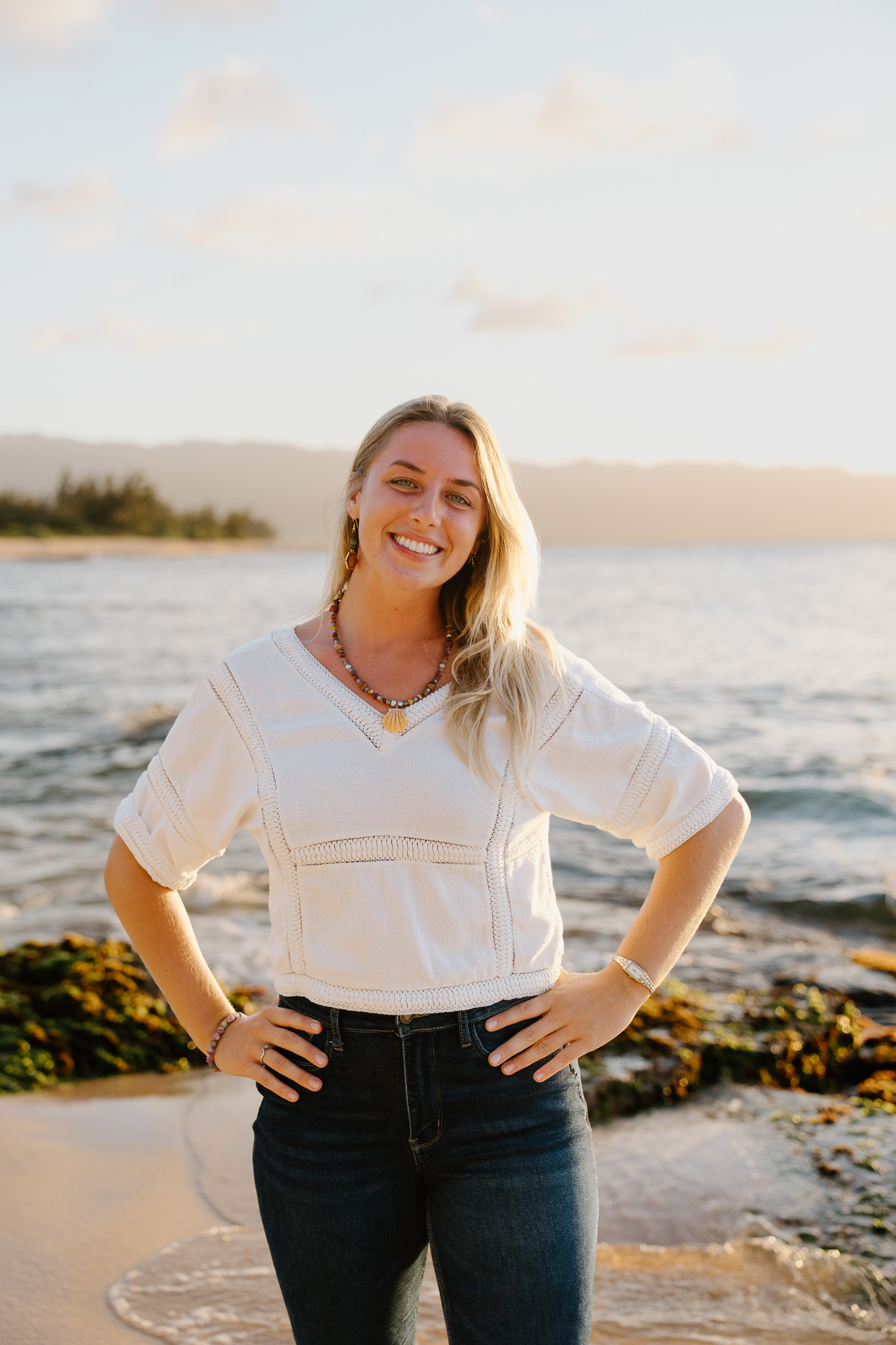 Lindsey on the beach at golden hour, in a cream top, hands on hips, smiling.